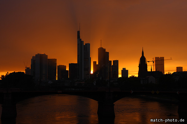 Frankfurter Hochhäuser vor orange leuchtendem Himmel mit Sonnenstrahlen.